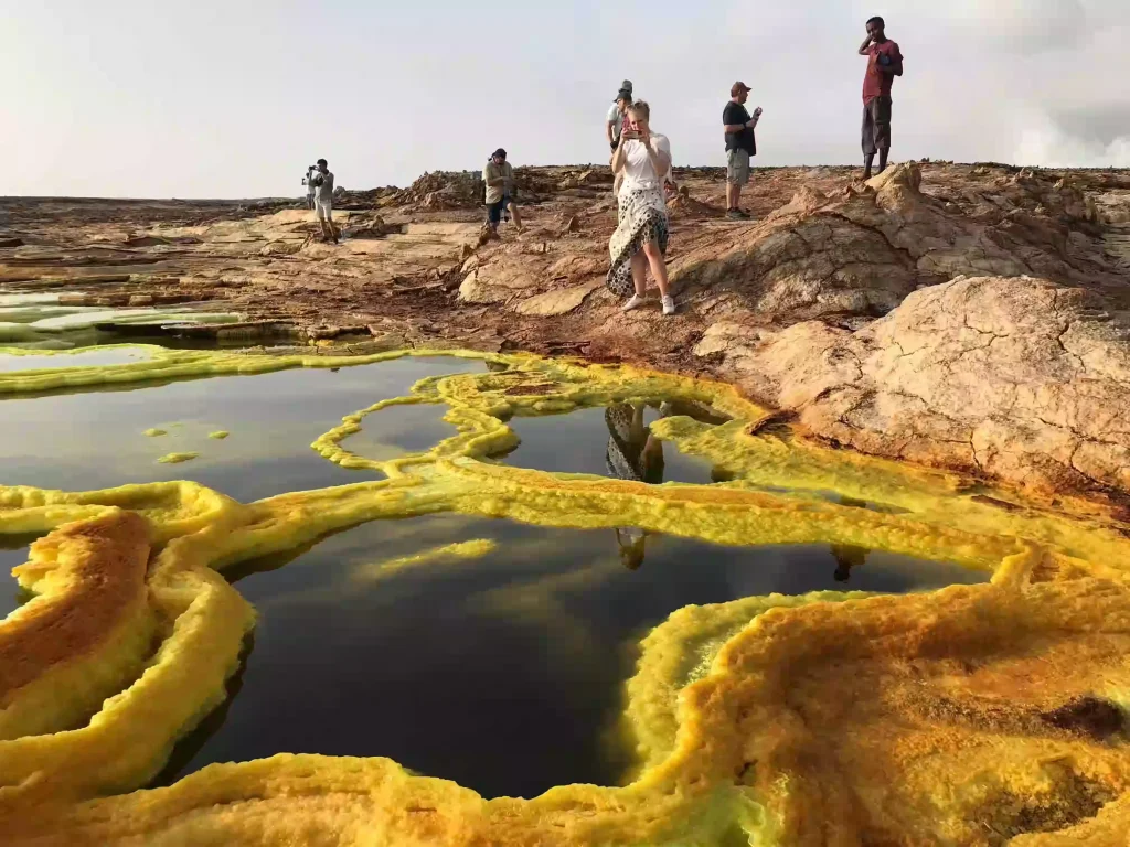 A photograph capturing the surreal landscape of Dallol in the Danakil Depression, Ethiopia, with our Tanian Ethiopia Tours clients in the frame. The image showcases the vibrant colors and otherworldly formations of Dallol's salt plains, hot springs, and mineral deposits. Clients, likely on a guided tour, are positioned to appreciate the vastness and unique beauty of the area. The Danakil's extreme environment is visually represented through the landscape's textures and colors.