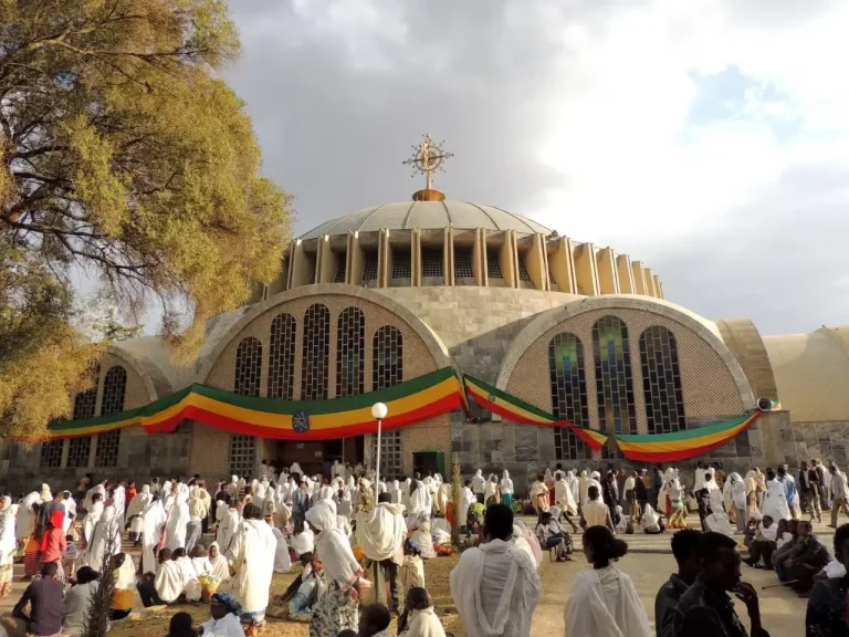 A wide shot of St. Mary of Zion Church in Axum, Ethiopia, showing the modern cathedral building with its large dome. Banners in the green, yellow, and red of the Ethiopian flag are draped around the church. A large crowd of people, many dressed in white, are gathered in front of the church, suggesting a religious service or celebration. The image captures a scene at this significant religious site in Axum.