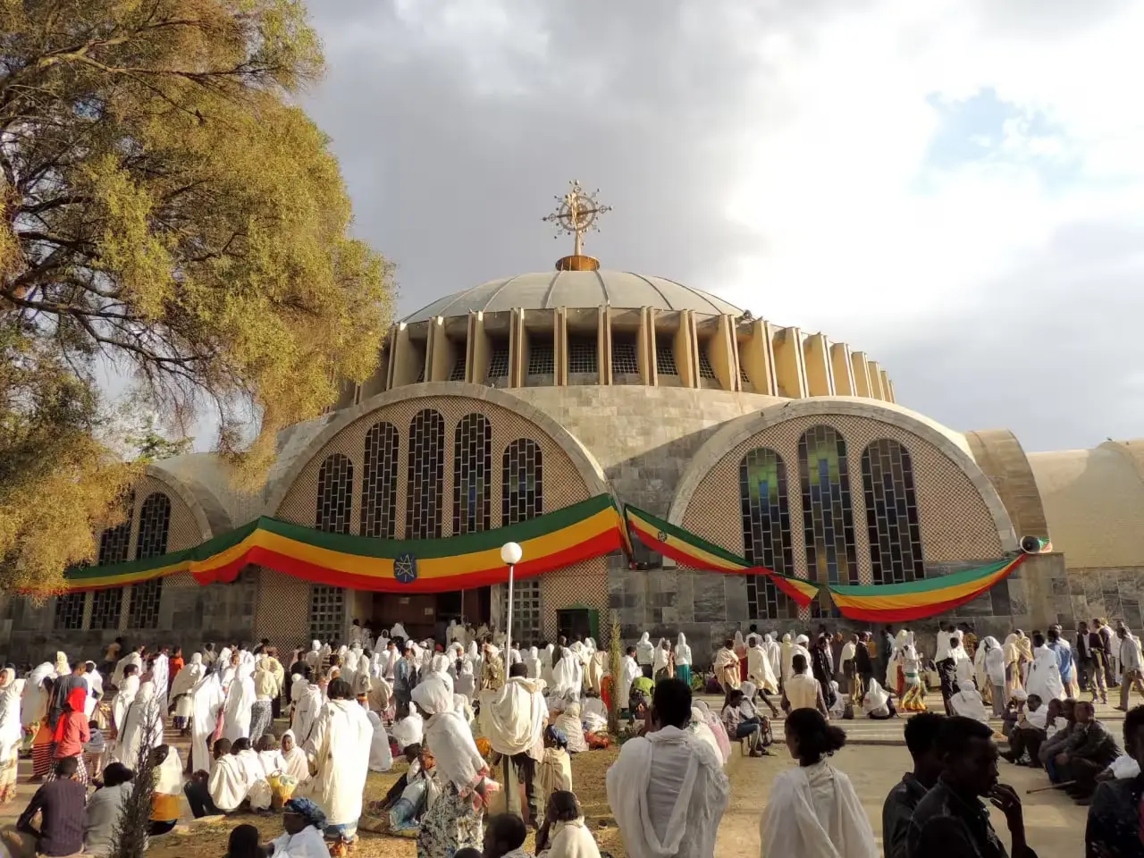 A wide shot of St. Mary of Zion Church in Axum, Ethiopia, showing the modern cathedral building with its large dome. Banners in the green, yellow, and red of the Ethiopian flag are draped around the church. A large crowd of people, many dressed in white, are gathered in front of the church, suggesting a religious service or celebration. The image captures a scene at this significant religious site in Axum.