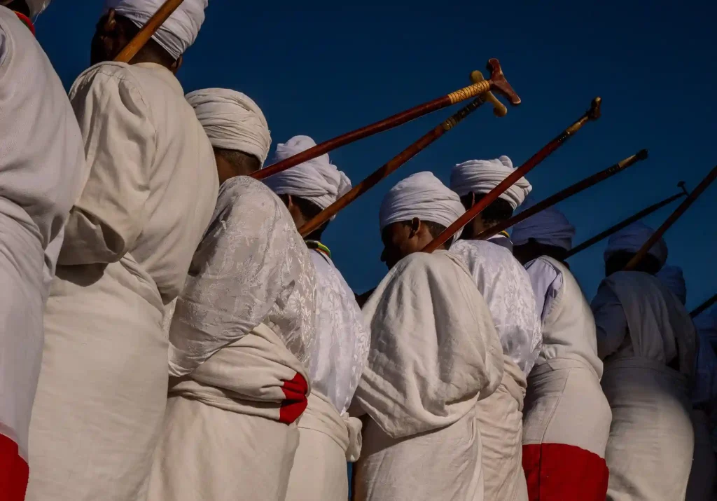 Close-up photograph showing a line of people dressed in white robes and head coverings, participating in a procession for Genna (Ethiopian Christmas) in Lalibela, Ethiopia. They are carrying wooden staffs.