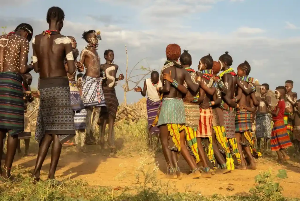 A wide shot captures a traditional circle dance performed by women of the Hamar tribe in Ethiopia. The women, distinguished by their ochre-clay styled hair and colorful patterned skirts, are linked arm-in-arm in a tight circle, dancing on dusty ground. Men in cloth wraps stand around the perimeter, observing. The background shows a dry, dusty landscape with sparse vegetation and a cloudy sky.