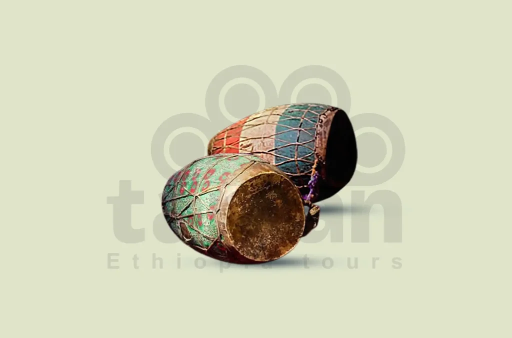 Studio shot photograph of two Kebero drums, traditional large, double-headed drums used in Ethiopian Orthodox Tewahedo Churches. The drums are decorated with colorful patterns and mesh-like netting. They are presented on a plain light background. The "Tanian Ethiopia tours" logo, watermarked in a light grey tone behind the drums.