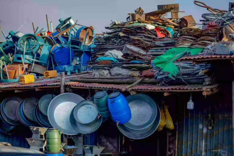 A vibrant and cluttered outdoor scene depicts a 'mercato,' likely a market in an African or Middle Eastern context. The foreground and midground are dominated by a corrugated metal roof structure overflowing with a diverse collection of metal items: pots, pans, buckets, chair frames, and various unidentified objects. Many items hang from the roof's edge, while others are piled high behind. The scene is brightly lit, and the overall impression is of a bustling marketplace focused on used and recycled goods. The atmosphere is energetic and suggests a place of trade and informal economy.