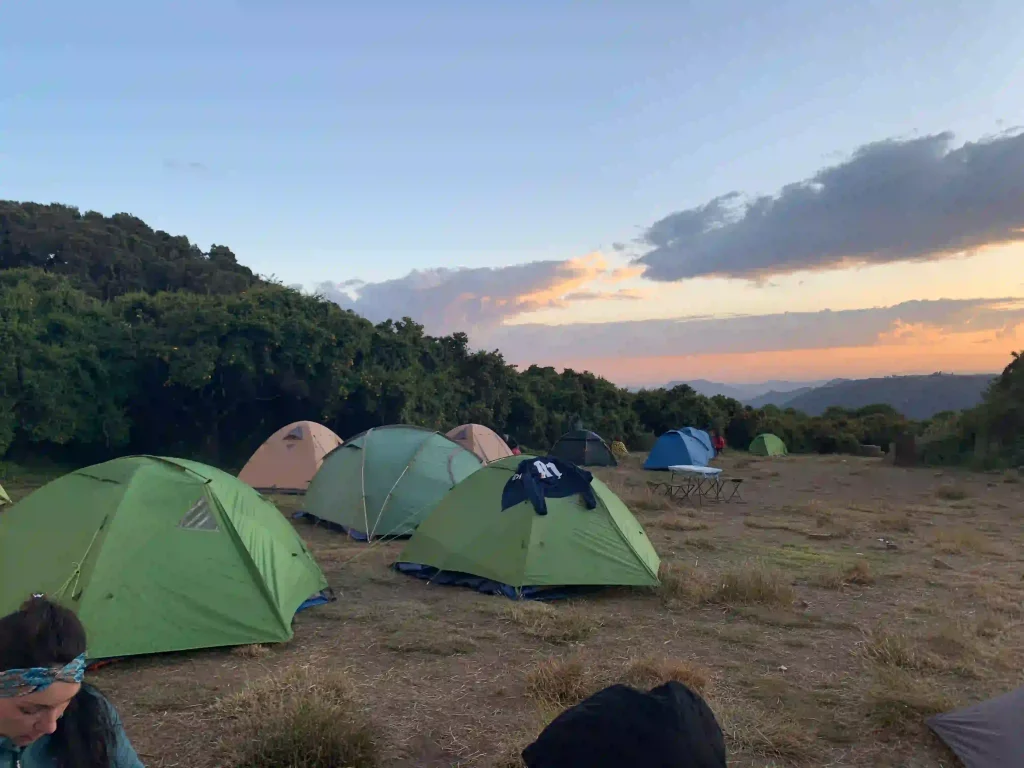 A wide shot shows a campsite in the Simien Mountains at dusk. Multiple tents, primarily green and beige, are pitched on a dry, grassy hillside. In the foreground, the back of a person wearing a dark beanie is visible. The background features a line of trees and a sunset sky with pink and orange hues.