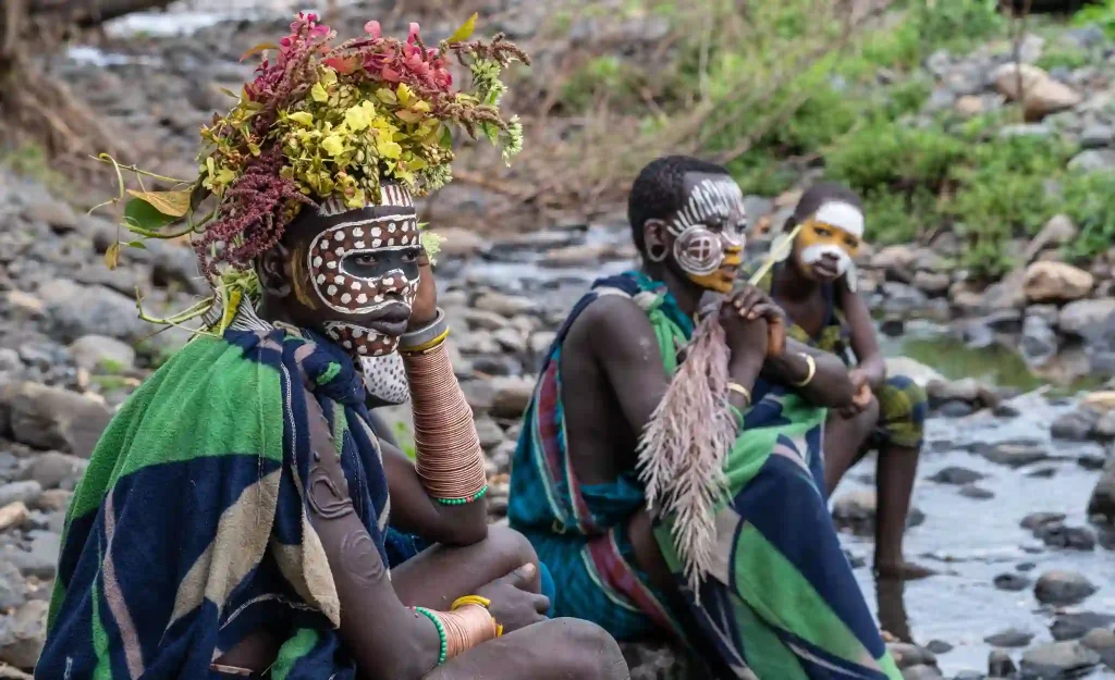 Three Surma women in traditional attire rest by a river, showcasing intricate facial paint designs and colorful patterned fabrics. The woman on the left is adorned with a vibrant flower headdress.