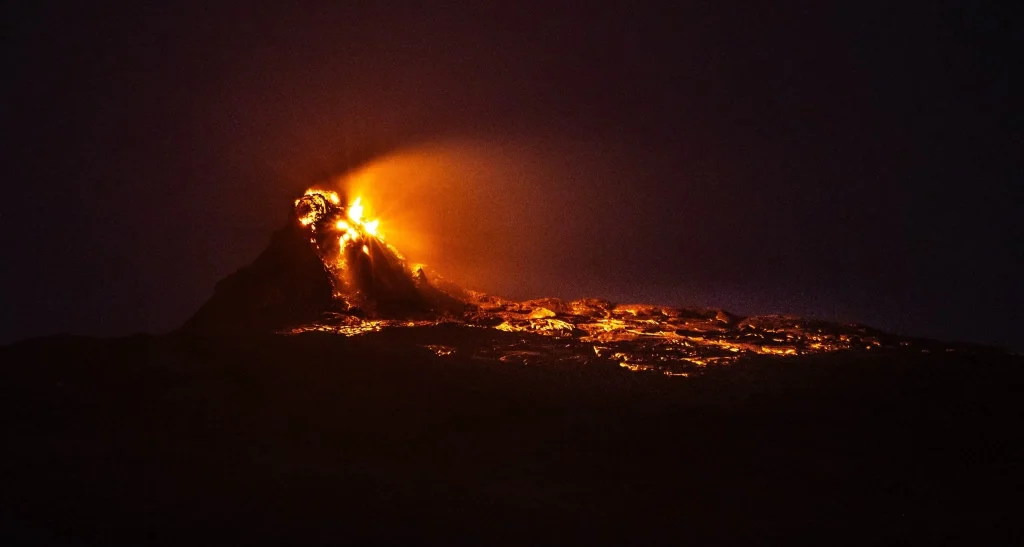 A nighttime photograph of Erta Ale volcano's lava lake in the Danakil Depression, Ethiopia, taken on a Tanian Ethiopia Tour. The image showcases the intensely bright orange glow of the lava lake within the volcanic crater, a signature feature of Erta Ale. The surrounding volcanic cone and landscape are silhouetted against the night sky, emphasizing the fiery spectacle. The mention of Tanian Ethiopia Tours suggests this is a promotional image, inviting viewers to experience this unique volcanic site with a guided tour.