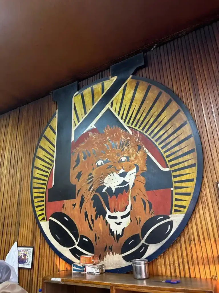 Photograph of the Tomoca Coffee logo. It features a stylized lion's head within a circular design with a large black letter "K". Coffee beans are visible beneath the lion.