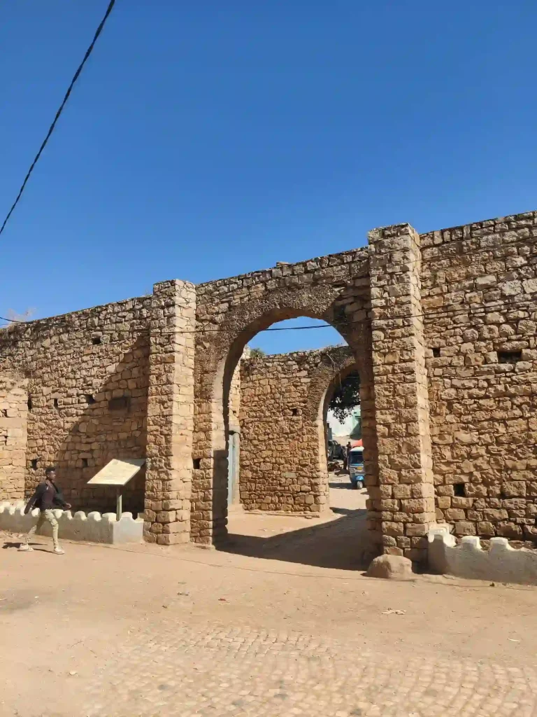 A section of the ancient stone walls of Harar Jugol, a UNESCO World Heritage Site in Ethiopia. The image shows an arched gateway leading into the city, with a man walking in the foreground.