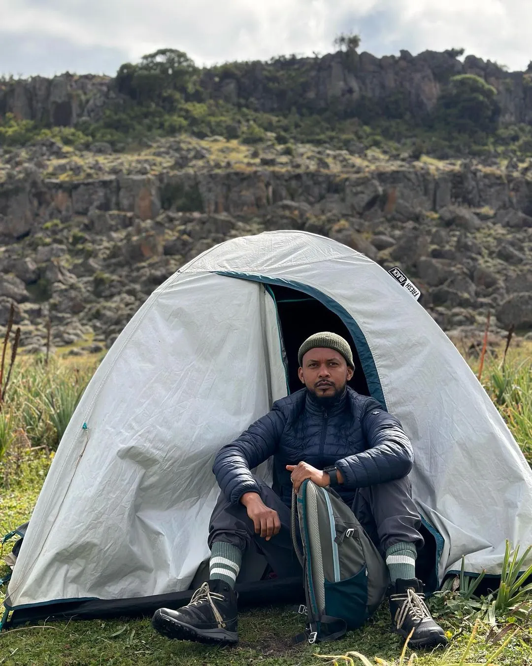 Nati, the owner of Tanian Ethiopia Tours, is pictured camping in front of a white tent in Bale Mountains National Park. He is seated with his backpack beside him, set against a backdrop of rocky hills and grassy terrain.