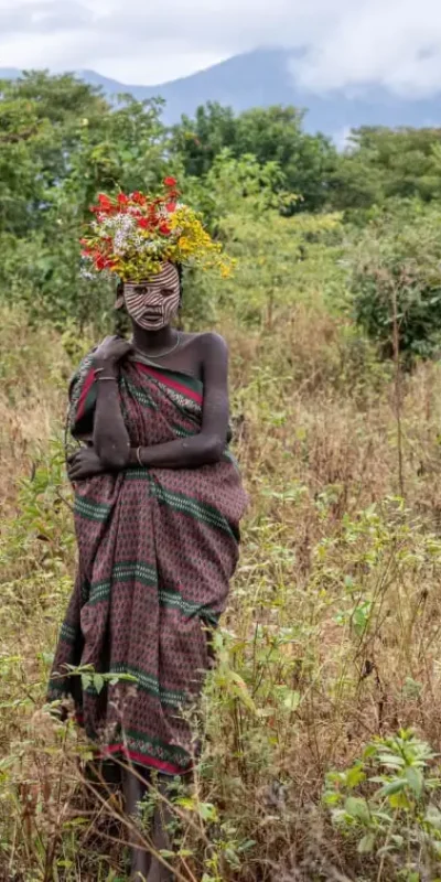 Medium shot photograph of a Surma woman standing in an outdoor setting of tall, dry grass and trees. She is facing forward, adorned with facial paint and a vibrant headdress of red, yellow, and white flowers. She wears a draped, patterned fabric and the misty blue mountains of Ethiopia are visible in the distance. This image captures the cultural adornment and natural beauty associated with Surma women.