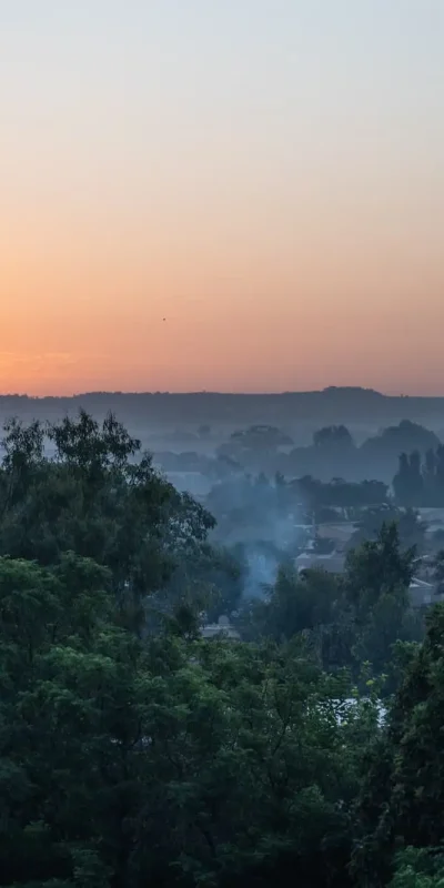 A scenic, wide shot captures the Bahir Dar cityscape during sunrise or sunset. The sky transitions from a warm orange and yellow at the horizon to a soft, hazy blue above. Lush green trees dominate the foreground and midground, partially obscuring rooftops and buildings. In the distance, a prominent, tall building rises above the skyline. The air appears misty, creating a soft, muted quality to the overall scene. The atmosphere is peaceful and serene, showcasing the beauty of Bahir Dar.
