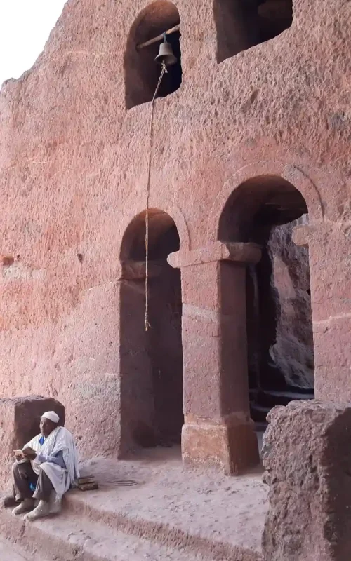 Exterior view of Bete Maryam (House of Mary) church in Lalibela, Ethiopia. This rock-hewn church is carved directly into the red volcanic rock. The image features arched doorways and windows, and a bell hanging from a rope. A person in traditional religious clothing is seated near the entrance, adding a human scale to the ancient structure. This photograph showcases Bete Maryam, one of the most famous churches of Lalibela.