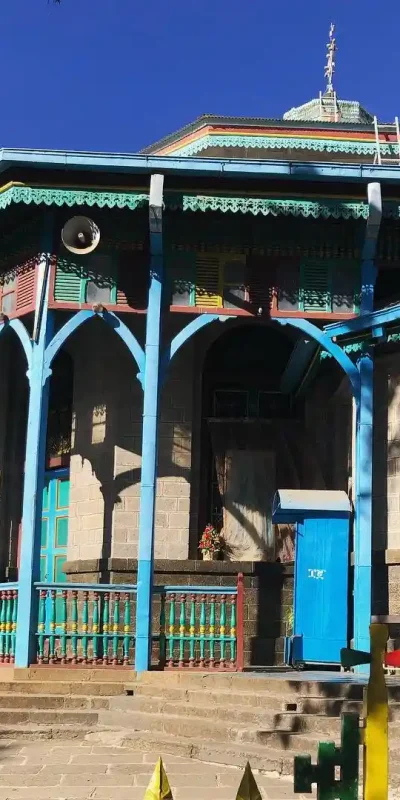 An image of Entoto Maryam Church, a historically significant church located on Entoto Hill in Addis Ababa, Ethiopia. The church's exterior features its characteristic bright colors – red, yellow, green, and blue – adorning its stone and corrugated iron structure. Entoto Maryam is associated with Emperor Menelik II and is a prominent religious and historical site in Addis Ababa.