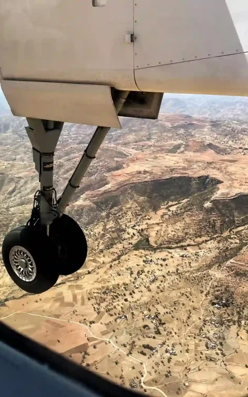 A photograph taken from an airplane window, showcasing the Ethiopian landscape with its varied terrain, including hills, valleys, and scattered vegetation. The airplane's landing gear is prominently visible in the foreground.