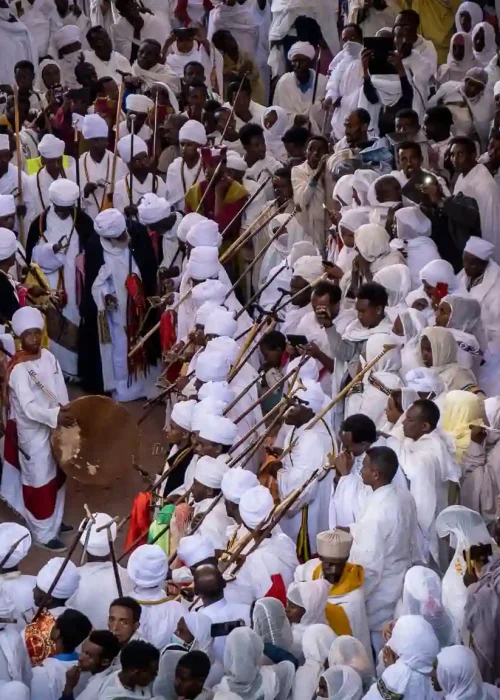 Ethiopian Orthodox priests in white robes stand on a hillside in Lalibela during the Genna celebration, responding to hymns and chanting from Bete Maryam Church. The image captures a moment of deep religious devotion and solidarity during Genna ceremony.
