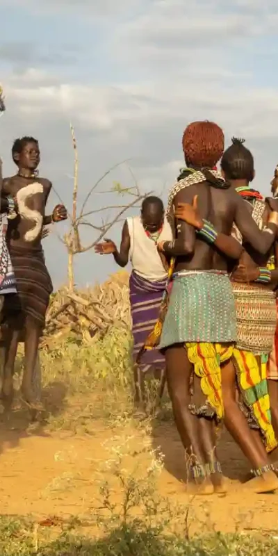 A wide shot captures a traditional circle dance performed by women of the Hamar tribe in Ethiopia. The women, distinguished by their ochre-clay styled hair and colorful patterned skirts, are linked arm-in-arm in a tight circle, dancing on dusty ground. Men in cloth wraps stand around the perimeter, observing. The background shows a dry, dusty landscape with sparse vegetation and a cloudy sky.