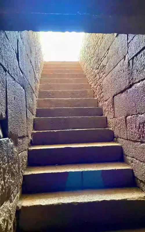 Low angle photograph of a stone staircase going upwards. The steps and walls are constructed from rough-hewn, weathered stone blocks. Bright sunlight streams in from an opening at the top of the stairs, illuminating the steps and creating strong shadows. This is within an ancient site in Axum, Ethiopia, possibly related to King Kaleb's tomb.