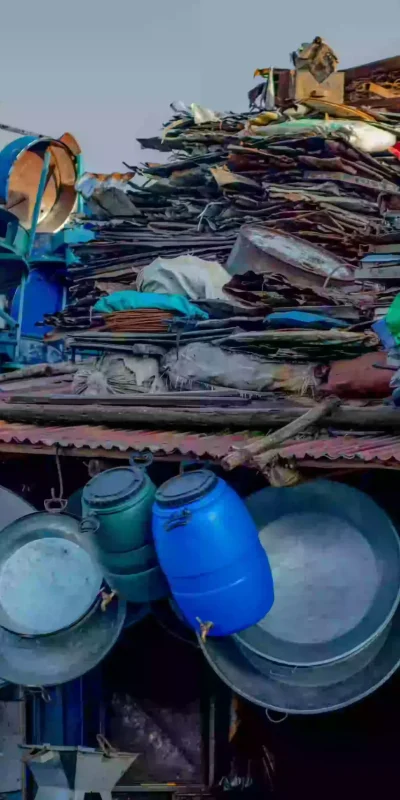 A vibrant and cluttered outdoor scene depicts a 'mercato,' likely a market in an African or Middle Eastern context. The foreground and midground are dominated by a corrugated metal roof structure overflowing with a diverse collection of metal items: pots, pans, buckets, chair frames, and various unidentified objects. Many items hang from the roof's edge, while others are piled high behind. The scene is brightly lit, and the overall impression is of a bustling marketplace focused on used and recycled goods. The atmosphere is energetic and suggests a place of trade and informal economy.