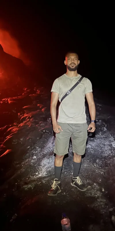 Nighttime photograph at Erta Ale volcano featuring Nati of Tanian Ethiopia Tours. The image contrasts the dark volcanic landscape and Nati's figure in the foreground with the intense red and orange glow of the active volcano in the background. An adventurous and dramatic scene.