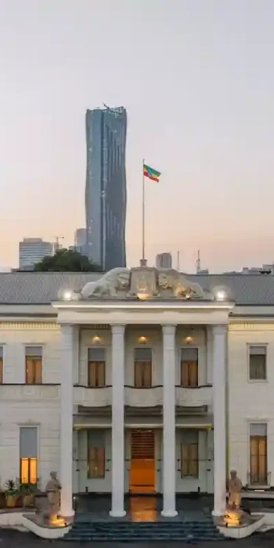 A ground-level, frontal photograph of the National Palace in Addis Ababa, Ethiopia, taken at dusk. Formerly the palace of Haile Selassie, now newly restored. The palace is a long, white building with a central portico featuring columns and lion statues. Ethiopian flags are prominently displayed on poles in front of the palace. Two tall palm trees flank the entrance. In the background, the modern skyline of Addis Ababa is visible against a soft, evening sky. The lighting is warm and highlights the architectural details of the palace.