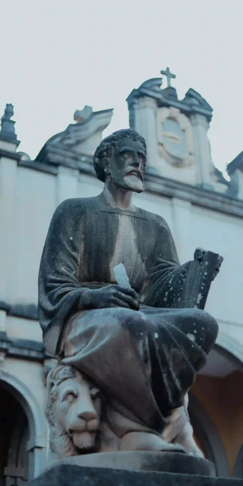 A statue is shown at Holy Trinity Cathedral in Addis Ababa, Ethiopia. The statue depicts a seated, bearded figure, possibly a saint or apostle, holding a book or scroll. A lion is sculpted at the base of the statue. The background reveals architectural details of the cathedral. This image captures a religious artwork within the grounds of Holy Trinity Cathedral, a prominent landmark in Addis Ababa.