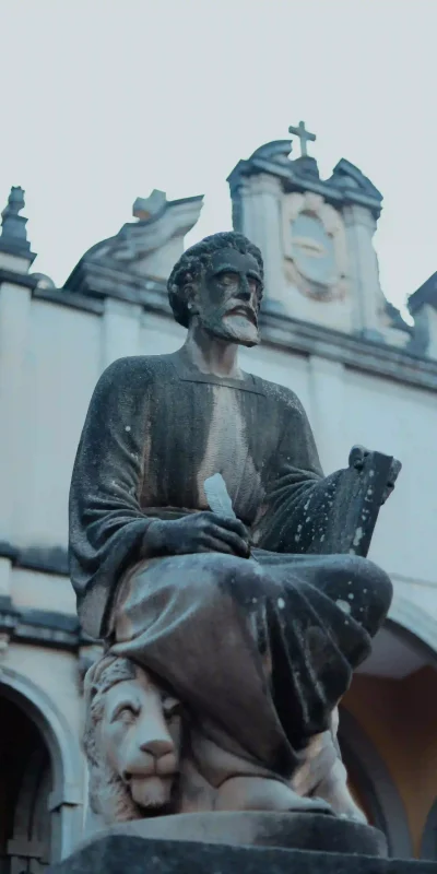 A statue is shown at Holy Trinity Cathedral in Addis Ababa, Ethiopia. The statue depicts a seated, bearded figure, possibly a saint or apostle, holding a book or scroll. A lion is sculpted at the base of the statue. The background reveals architectural details of the cathedral. This image captures a religious artwork within the grounds of Holy Trinity Cathedral, a prominent landmark in Addis Ababa.