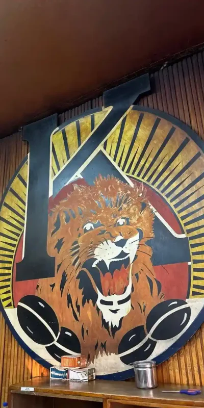 Photograph of the Tomoca Coffee logo. It features a stylized lion's head within a circular design with a large black letter "K". Coffee beans are visible beneath the lion.