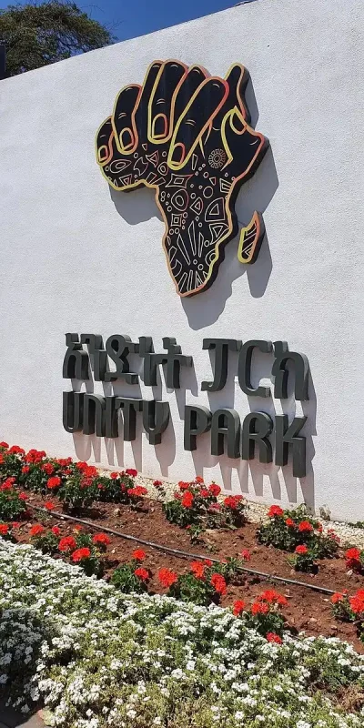 The entrance to Unity Park in Addis Ababa, Ethiopia is marked by a prominent white wall. Affixed to the wall is a 3D sign featuring a stylized black and gold hand holding the continent of Africa. The African continent is decorated with intricate patterns. Below the hand, in large, stylized grey lettering, are two lines of text. In front of the wall, neatly planted rows of red and white flowers create a colorful border. A bright blue sky is visible above. The overall impression is of a grand and welcoming entrance to Unity Park, a significant landmark in Addis Ababa.
