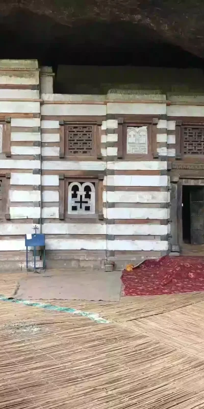 Photograph of the distinctive exterior of Yemrehana Krestos Church in Lalibela, Ethiopia, known for its unique striped facade of alternating dark wood and white stone. Intricate wooden windows and a rocky overhang are also visible.